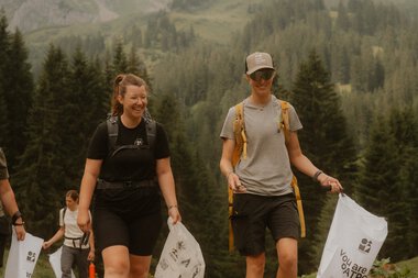 Zwei junge Menschen wandern auf einem Waldweg in den Bergen, umgeben von Tannen und hohen Gipfeln, bei einem Umwelt-Cleanup-Event. | © Patron e.V. • Fotograf: Michelle Meitzner