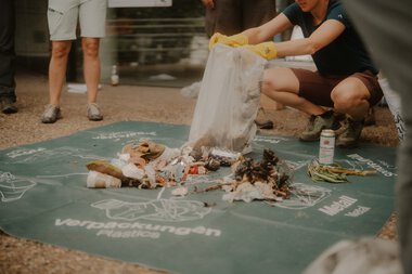 Personen sammeln Müll beim Cleanup-Event, um die Umwelt zu schützen, auf einer Matte mit Müll, Handschuhen und Recycling-Tüte. | © Patron e.V. • Fotograf: Michelle Meitzner