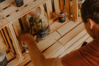 Mensch bei einer Umweltaktion, der verschiedene Glasbehälter mit Müll sortiert. Die Gegenstände sind auf einem Holzregal aus Holzlatten abgelegt. | © Patron e.V. • Fotograf: Michelle Meitzner