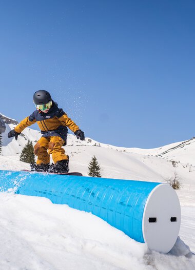 Ein Kind beim Snowboarden im verschneiten Gebirge mit blauer Himmel, das auf einem gebogenen, blauen Rail fährt.