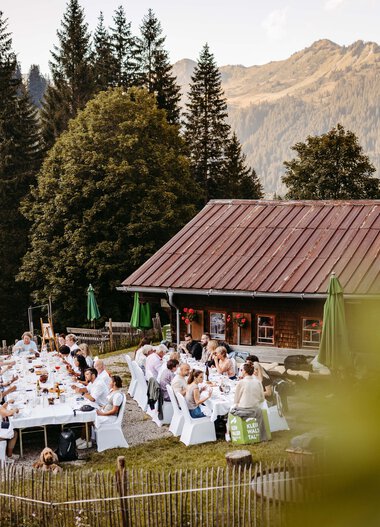 Eine große Gruppe sitzt an festlich gedeckten Tischen vor einer Berghütte in alpiner Landschaft. | © Kleinwalsertal Tourismus eGen • Fotograf: Sascha Gast